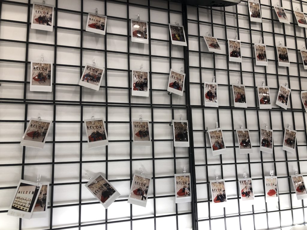 Ein schwarzes Gitter vor einer weißen Wand, an dem Polaroidfotos hängen