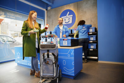 Eine Frau mit langen Haaren und einem Einkaufstrolley an einer Theke einer Abholstation, wo sie von einem Mitarbeiter bedient wird