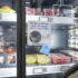 Ein Kühlregal mit Glastür mit Lebensmitteln darin, eine Hand hält ein Smartphone davor