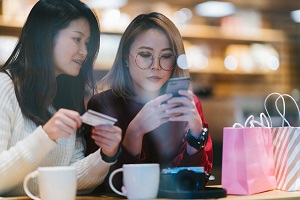 Two female Asian friends are shopping online while using a credit card and a smartphone in a cafe.; copyright: Worldpay by FIS GmbH