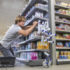 Ein Mann im Supermarkt kniet vor einem Regal und Deosprühflaschen fallen an der Seite aus dem Regal