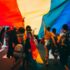Many people celebrate in the street with arms raised under a huge rainbow flag