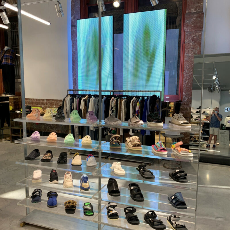 A shoe rack in a store, behind it large screens on the wall