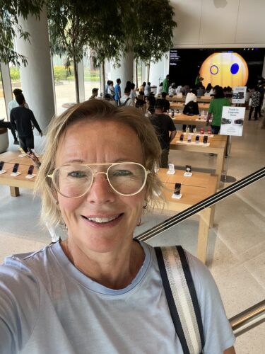 A woman with blonde hair and glasses in the foreground of the selfie, behind it a view of the Apple store