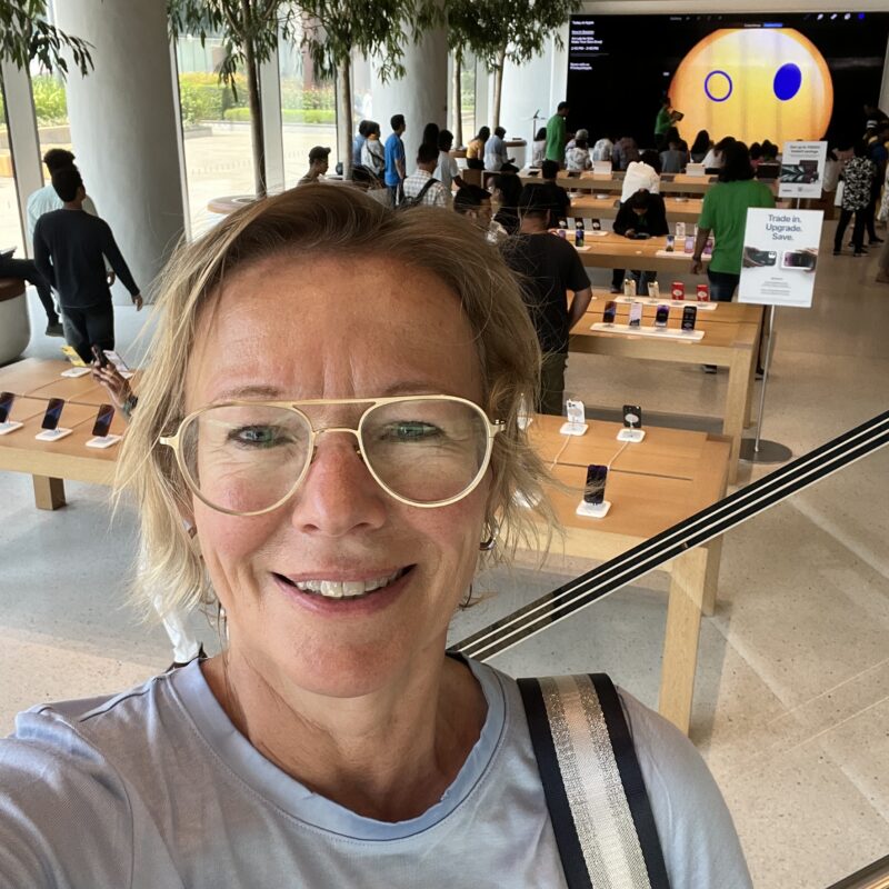 Eine Frau mit blonden Haaren und Brille im Vordergrund des Selfies, dahinter ein Blick in den Apple-Store